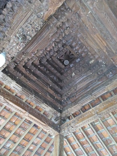 An upward-angled view captures a richly carved, dark-stained wooden ceiling within a structure in Dulce Nombre de Jesús, Costa Rica. The ceiling features a complex coffered or pyramidal design composed of multiple layers of wooden beams and panels, all intricately carved with floral, geometric, and scrollwork motifs. A central square opening is present at the apex, containing a small, light-colored circular object, likely a vent or fixture. White specks, consistent with dust or mildew, are visible across the dark wood surfaces. A partial view of a white, opaque light fixture is visible in the upper-left quadrant. On the lower sides of the image, sections of a sloped roof constructed from overlapping terracotta tiles and supported by wooden rafters are visible. Heavy, carved wooden beams and a structural pillar are also visible, connecting the roofline to the ornate ceiling.