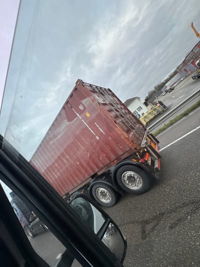 The image depicts an outdoor scene under an overcast sky in Fegersheim, France. A dark red, weathered shipping container, marked "TCSU 086989 2" and "42G1", is observed from the right side. It is loaded onto a multi-axle semi-trailer, visible with two axles featuring dual wheels and reflective hubcaps. The trailer's rear lighting assembly is illuminated. The vehicle is situated on an asphalt road with a prominent white lane line and visible tire tracks on the wet surface. The photograph is captured from within another vehicle, with portions of the dashboard, window frame, and rearview mirror visible in the foreground. The rearview mirror reflects part of a truck behind the camera's position. In the background, a light-colored building with a brown roof stands near a yellow barrier, adjacent to industrial-style buildings and leafless trees. A segment of a yellow construction crane arm is visible on the far right.