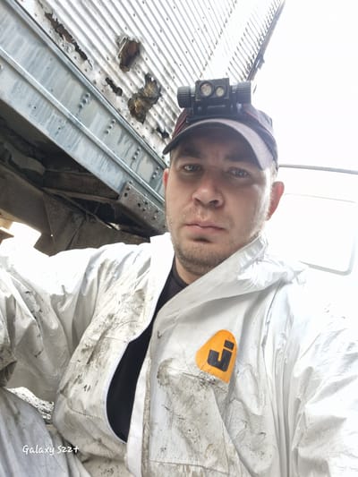 An outdoor, close-up, selfie-style photograph shows a man wearing a white disposable coverall suit, a black t-shirt, and a baseball cap. He is wearing a headlamp attached to his cap. The man's face is visible, and he appears to be looking at the camera. The coverall suit is stained with dirt and grime. The background shows a section of a corrugated metal structure that appears to be damaged and rusted. The lighting suggests it is daytime. The image is tagged "Galaxy S22+". The specific location in Zheleznogorsk-Ilimsky, Russia, is not determinable from the visual information provided in the image.