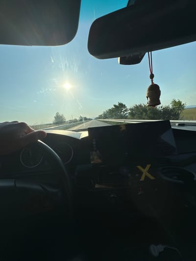 A first-person perspective photograph captures the interior of a moving vehicle looking forward through the windshield, on a sunny day in Mogila, Bulgaria. The sun, a bright white-yellow orb, is low in the sky, positioned slightly left of center, causing significant glare and visible streaks on the windshield.
In the foreground, a human left hand grips the top-left portion of a black, three-spoke steering wheel. Behind the steering wheel, two dark-faced analog gauges are partially visible on the dashboard. Above, a light-colored sun visor is partially visible on the upper left, while the underside of a rearview mirror assembly is prominent in the upper right. A small, wooden or dark-toned Buddha head charm, attached by a red cord, hangs from the rearview mirror.
To the right of the steering wheel, a large, dark, rectangular screen, possibly an infotainment system or tablet, is mounted on the dashboard. Below this screen, a yellow 'X' symbol or sticker is affixed to the dashboard surface. Further to the right, a circular compartment is present. In the bottom right corner of the frame, a white cable and a small white object are visible on the floor or lower console.
Through the windshield, a two-lane asphalt road with white markings extends into the distance, flanked by green vegetation and trees. Utility poles and power lines parallel the road on the left. Distant hills, covered in vegetation, are discernible under a clear blue sky with some atmospheric haze.