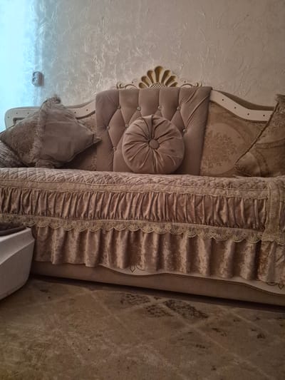 The image shows a close-up of an ornate, upholstered sofa in a room. The sofa is a light brown or beige color and features tufted detailing on the back cushions, adorned with clear buttons. There are several decorative pillows on the sofa, including a round, tufted pillow and a square pillow with a floral pattern. The sofa also has a decorative skirt with ruffles and lace trim. The wall behind the sofa has a textured finish and a light fixture is visible on the left side. The floor in front of the sofa is covered with a textured carpet. No people or specific city landmarks are visible, making it impossible to determine the exact location in Қанабек, Kazakhstan.