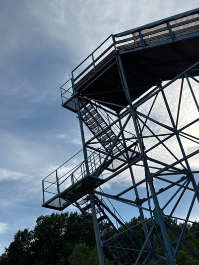 The image displays a large, multi-level metal observation tower, located in the city of Svetloe, Russia, captured from a low, upward perspective. The tower's structure consists of dark grey metallic beams forming a complex, crisscrossing lattice pattern that supports at least two distinct platforms. Each platform features wooden decking and is secured by metal railings. Metal staircases with grated treads and handrails provide vertical access between these levels. The background is dominated by a partly cloudy sky, showing patches of muted blue and grey interspersed with diffuse white, wispy clouds. Sunlight, emanating from the upper right, partially illuminates the metallic framework and the cloud formations. Below the visible structure, a dense expanse of dark green trees delineates the horizon, suggesting a forested or heavily vegetated environment. No people are present within the frame.