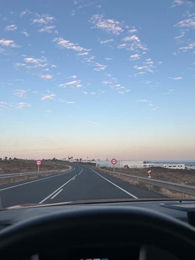 The image shows a road in Tías, Spain, with a blue sky and scattered clouds above. The road curves to the right, with guardrails along the side. There are two red and white circular traffic signs on the left side of the road, indicating no overtaking. In the distance, there are some buildings and palm trees. The photo appears to be taken from inside a car, with the dashboard visible in the foreground. The overall impression is of a scenic drive in a coastal area.