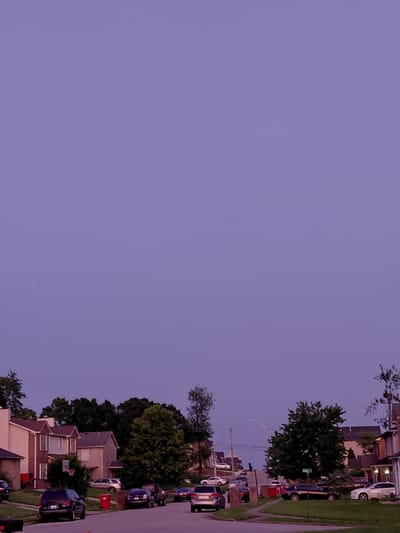 The image captures a suburban street in Nicholasville, USA, at dusk. The sky is a deep, hazy purple, suggesting twilight or perhaps an impending storm. The foreground features a street lined with parked cars, including a black SUV, a dark sedan, and a silver sedan, among others. Houses with visible architectural details like pitched roofs and windows are scattered along the street, bordered by green lawns and mature trees. A basketball hoop is visible attached to one of the houses. The overall impression is a quiet residential neighborhood transitioning into evening.