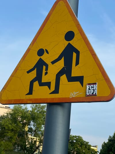 The image shows a yellow triangular traffic sign with a black border, mounted on a grey metal pole. The sign depicts two black silhouetted figures, one adult and one child, running from left to right. The sign is weathered with visible cracks in the yellow surface. On the lower right side of the sign, there is a sticker with the text "FCK UPA" in white and red lettering against a black background, and a small drawing in white. The background is a clear blue sky with some faint clouds. Below the sign, green trees and the upper parts of buildings are visible, suggesting an urban or suburban outdoor setting. The overall scene appears to be a street or roadside in the city of Oława, Poland.
