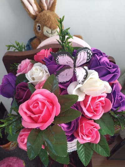 This media file features a close-up shot of a decorative arrangement of artificial roses and greenery, possibly a bouquet or a centerpiece. The roses are in various shades of pink, white, and deep purple. A decorative butterfly with pink wings and black polka dots is positioned prominently among the flowers. In the background, a plush toy rabbit with long ears and a blue jacket is visible, peeking over the edge of what appears to be a wooden shelf or basket. The overall impression is of a cheerful and whimsical display, likely intended for decoration. The lighting suggests an indoor setting, possibly during daylight hours. There is no visible text in the image. The location context of Darlington, United Kingdom, does not provide specific visual cues within the image itself.