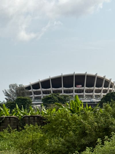 The image presents a distant, elevated view of the National Arts Theatre located in Ebute-Metta, Lagos, Nigeria. The prominent structure is a large, curvilinear building featuring a dark, curved roof supported by numerous light-colored, vertical concrete ribs or pillars. Several levels with rounded openings are visible between these supports. A single red banner or sign is affixed to the building's facade on the right side of the visible structure. The lower section of the building is significantly obscured by dense, verdant vegetation, which dominates the foreground and midground. This foliage includes various shrubs, trees, and broad-leafed plants, some resembling banana leaves, which rise from a concrete barrier in the immediate foreground. To the left of the main structure, a tall, slender tree with sparse, light-colored foliage is visible. The sky above is largely overcast with scattered light grey clouds. No individuals, specific actions, or interactions are discernible within the frame.