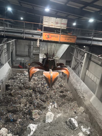 The image shows an industrial setting with a large metal claw excavator digging into a massive pile of mixed waste. The waste appears to consist of various materials, including plastics and other debris, suggesting a recycling or waste management facility. The excavator is positioned to scoop up the waste from a concrete containment area. In the background, structural elements of the building, including support beams and lighting fixtures, are visible. The presence of "IMO-CZ Automatizace" and "Товароподемност 10 500 kg" on a large sign suggests the crane is capable of lifting 10,500 kg. The specific location is Beli Izvor, Bulgaria.