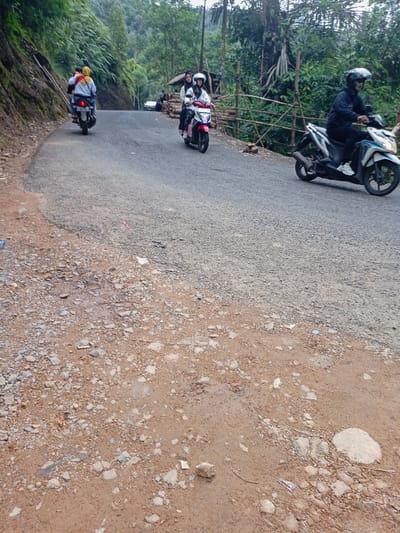 The image shows a paved road in a rural area of Cisewu, Indonesia (latitude: -7.411, longitude: 107.542).  There are three motorcycles traveling on the road. The motorcycle in the foreground is being driven by a man wearing a helmet and black clothing. The motorcycle in the middle is being driven by a woman wearing a hijab and light-colored clothing, with a passenger behind her. The motorcycle in the background is being driven by a man wearing a helmet and light-colored clothing. All motorcycles are traveling in the same direction, and there are no interactions between the drivers. The road is lined with a wooden fence on the right side and a rocky embankment on the left. The embankment is covered in loose rocks and dirt. The surrounding area is wooded with trees on both sides of the road. The sky is cloudy and overcast.
