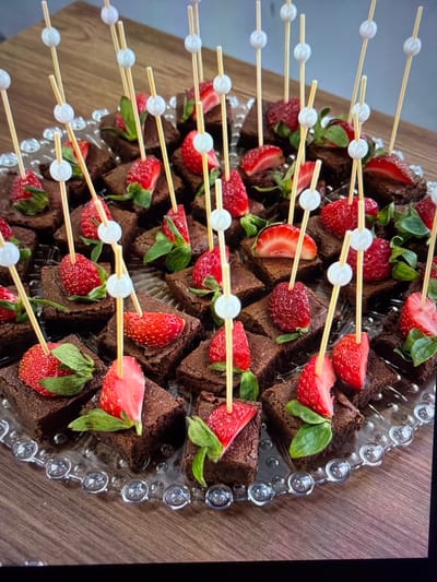 This image captures a close-up, high-angle view of a dessert presentation featuring numerous individual portions of brownies and fresh strawberries. The brownies are dark brown, appearing rich and moist, and are cut into small, irregular squares. Each serving is adorned with a bright red strawberry, many of which retain their vibrant green leafy caps, indicating freshness.

Thin wooden skewers, each featuring a small white spherical bead near its tip, are strategically inserted into many of the brownie and strawberry combinations. These skewers are arranged in neat, parallel lines extending diagonally across the frame, suggesting an organized and accessible display.

The desserts are arranged on a clear glass platter, which has a distinctive textured rim made up of a series of small, transparent glass spheres. The platter rests on a warm, polished wooden surface, likely a table, which is visible in the background and extending along the edges of the frame. The lighting is even, suggesting an indoor setting. This arrangement appears to be a prepared dessert spread, ideal for a party, gathering, or special event, potentially taking place in Ros Na Rí, Ireland. No people are visible, and there is no discernible text in the image.