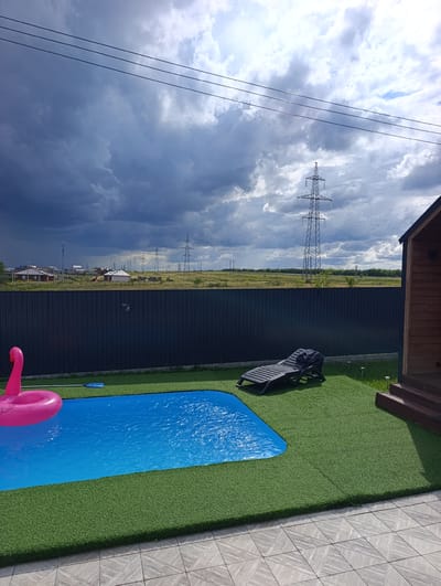 The image shows a backyard with a swimming pool and artificial grass. A pink inflatable flamingo floats in the bright blue water of the pool. A dark plastic lounge chair is placed on the grass near the pool, with a dark towel draped over it. To the right of the pool area is a small wooden structure with stairs leading up to it. The backyard is bordered by a dark fence. In the background, there is a field with some houses and numerous tall power line towers stretching across the landscape. The sky is filled with dramatic, dark storm clouds, suggesting an impending storm or the aftermath of one. The lighting indicates it is daytime, possibly late afternoon, given the contrast between the bright pool and the dark sky. The overall scene depicts a leisure space that is about to be interrupted by inclement weather. There is no visible text in the image.