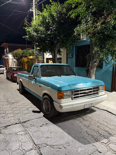 A blue and white pickup truck is parked diagonally on a brick-paved street. It is beside a teal building with a tree obscuring part of the view. Street lights are visible on the left. Other cars are parked behind the pickup. The scene is well-lit, and the street appears to be in a residential area.