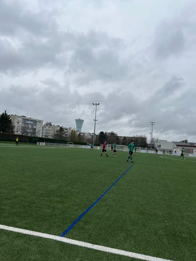 The image displays an outdoor artificial turf soccer field located at the Dezavelle stadium area in Montigny-lès-Metz, France, identified by the distinct Château d'eau de Montigny-lès-Metz - La Corchade water tower prominently visible in the background. The sky is entirely overcast, casting a muted light.
Approximately twelve individuals are visible on the field. Ten are soccer players, with at least six wearing green uniforms and at least three wearing red uniforms. One individual in a yellow top and dark bottoms stands near the left sideline. A player in a green uniform walks diagonally across the mid-foreground from right to left. Other players are distributed across the field, standing or moving.
The field is marked with white lines, including one in the immediate foreground, and a diagonal blue line extending towards the center. Two white soccer goalposts are positioned in the mid-ground. Tall stadium floodlight poles with multiple lamps are visible, two of which stand prominently in the mid-ground. In the background, behind the field's perimeter fencing, several multi-story apartment buildings and numerous trees with sparse foliage are present beneath the light blue water tower.