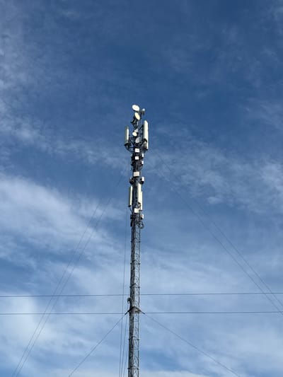 A tall, metallic telecommunications tower stands against a bright blue sky with scattered white clouds. The tower is segmented and appears to be a lattice structure, supporting numerous antennas and equipment at various heights. Several wires extend from the tower, disappearing out of frame. The image is taken from a low angle, looking up at the tower. The location is not specified.
