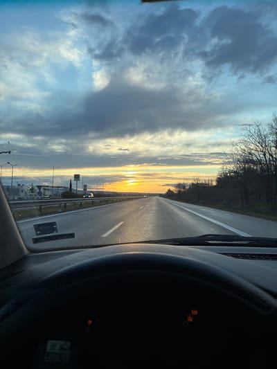 A view from inside a vehicle looking out onto a highway at sunset. The sky is partly cloudy with the sun setting on the horizon, creating a bright orange and yellow glow. On the right side of the road, there are trees. On the left side of the road, there is a gas station and traffic lights and traffic. The foreground of the image includes the interior of the vehicle, notably the dashboard and the steering wheel. The location is on a highway, possibly near Momkovo, Bulgaria.