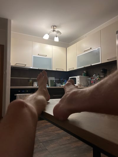 The image shows a first-person perspective of a person's bare feet resting on a table in a kitchen. The kitchen has cream-colored cabinets, dark countertops, and a tiled floor. There are various kitchen appliances visible, including a microwave, a coffee maker, and a refrigerator. The lighting suggests it is nighttime or indoors with artificial light. There is no specific indication of the location being in Rumia, Poland; this detail is an external assertion.