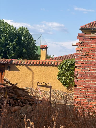 The image depicts an outdoor scene in Arcahueja, Spain, on a clear day with a blue sky dotted with wispy clouds. The foreground is dominated by dry, brown vegetation. In the midground, a textured yellow wall forms a prominent feature, topped by a terracotta tiled roof. A section of a red brick wall, partially constructed and with visible hollow bricks, stands to the right, partially obscuring the view of the yellow wall and roof. A chimney with a terracotta cap rises above the yellow wall. To the left of the yellow wall, a dark wooden door is visible. A pile of discarded wooden planks and rubble sits in front of the yellow and stone-patterned lower wall. Green ivy climbs on the yellow wall near the brick structure. In the background, a lush green tree with dense foliage is visible, and a chain-link fence with a green surface behind it suggests a recreational area, possibly a tennis court. A metal pole with a curved top extends from the rubble pile, and a small red and white flag is attached to it. A small light fixture is mounted on the yellow wall. The overall impression is one of a partially maintained or under-construction property.