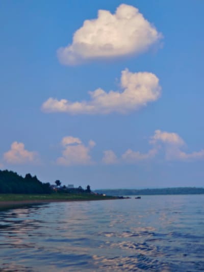 A calm body of water is in the foreground of the image, with a shoreline to the left featuring a line of trees and some distant buildings. The sky above is blue and clear, with a few scattered white clouds. The image is taken from a low angle, suggesting the vantage point is from a boat or the water's edge. The overall scene is tranquil and depicts a natural landscape. There are no people or animals visible in the image. The location in the city of Погодаево, Russia, is not explicitly indicated within the visual elements of the photograph itself.