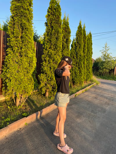 A young person wearing a black t-shirt, denim shorts, a black baseball cap, and pink Crocs stands on an asphalt road in what appears to be a suburban area, possibly in the city of Pushkino, Russia. The person is partially obscured by their hand held up to their face. They are positioned in front of a row of tall, green evergreen trees and a brown wooden fence. The sun is low in the sky, casting long shadows on the road and illuminating the scene with a warm, golden light. The environment suggests a residential neighborhood with clear signage of typical Russian suburban architecture.