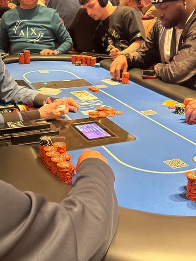 The image documents an indoor poker game at a blue felt table in Winchester, United States. Five individuals are visibly seated around the table, engaged in gameplay.
The blue felt poker table features light blue and white outlines for player positions and betting areas, with dark padded armrests. Embedded in the table is a small electronic display showing purple-hued text and numbers, likely game statistics. Several small white QR code-like squares are printed on the felt.
Poker chips are prominent. Large stacks of predominantly orange chips, with some black, white, and green chips mixed in, are positioned in front of players. Loose orange chips are scattered in the central betting area. Playing cards are visible on the table; some are face-up community cards (King of Spades, 10 of Spades, 8 of Spades, Queen of Clubs, 2 of Hearts, Ace of Diamonds), while others are face-down. One set of light-skinned hands, wearing a blue long-sleeved shirt, holds a stack of cards, indicating a dealer or a player handling their hand.
From left to right:
1. A person in a teal long-sleeved sweatshirt with an "ARMANI EXCHANGE" logo and a watch on their left wrist sits behind a stack of orange chips.
2. Next, a young man with dark hair wears black over-ear headphones and a dark t-shirt with a light graphic. He has a distinctive tattoo on his right forearm and is focused downwards at the table, with stacks of orange chips in front of him.
3. To his right, a dark-skinned man with a black bandana and a gold chain wears a black long-sleeved jacket. A red-cased smartphone rests on the table in front of him, beside his stacks of orange and multi-colored chips.
4. In the foreground on the far left, an arm in a dark long-sleeved shirt is visible next to a substantial stack of orange and multi-colored chips.
The scene is brightly illuminated by artificial light. The background is blurred, showing indistinct figures and warm-toned patterned wall details, consistent with a casino or gaming establishment.