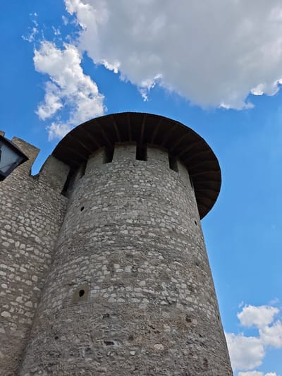 A low-angle shot captures the weathered stone exterior of a round fortress tower against a vibrant blue sky dotted with fluffy white clouds. The tower, constructed from rough-hewn stones of varying sizes and shades of gray, features small rectangular openings near its top, suggesting defensive windows or embrasures. A wooden roof, supported by visible beams, crowns the structure, its edges serrated. To the left, a portion of an adjacent stone wall extends upwards, also showing signs of age and rough construction. The overall impression is of an ancient defensive structure, possibly part of the Soroca Fortress in Moldova, emphasizing its robust construction and integration with the natural sky.