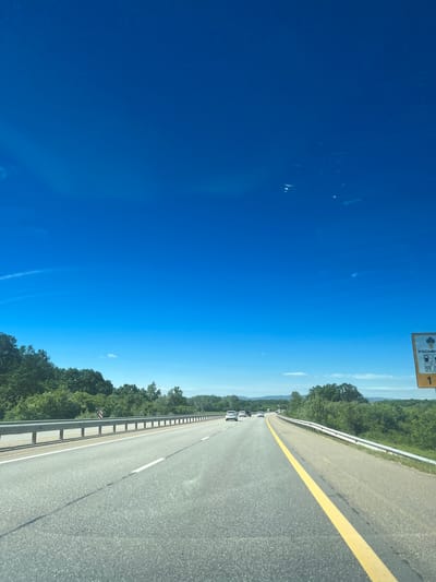 The image depicts a view from a vehicle traveling on a highway in Saratovskaya Oblast, Russia. The scene captures a straight section of the road with two lanes in each direction. A solid yellow line divides the lanes, and a metal guardrail runs along the roadside. The road is lined with trees. Several vehicles are visible in the distance on the same lane. The sky is clear and blue. A highway sign can be seen in the top left of the frame.
