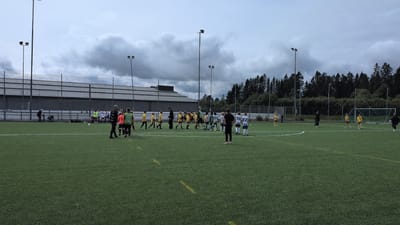 This is a horizontal, eye-level shot of a youth soccer game on an artificial turf field in Brunflo, Sweden. The sky is overcast with dramatic, dark clouds, suggesting either early morning or late afternoon under inclement weather. The field is green with white markings for the game. A group of children in yellow and green uniforms are gathered on the field, some interacting with adults in black attire, likely coaches or referees. In the background, a fence with netting surrounds the field, and beyond that, a building with a grey facade and a row of dark green coniferous trees line the horizon. Tall light poles with lamps are visible on either side of the field. The overall mood appears to be serious, with the children focused on the game. There is no visible text in the image.