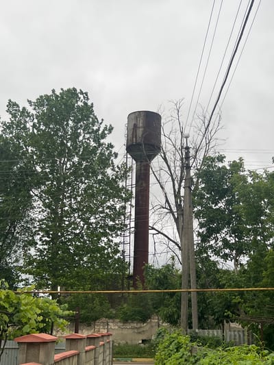 An old, rusty water tower stands tall amidst lush green trees and vegetation under a cloudy sky. The tower, made of metal, shows signs of wear and tear, with a ladder running up its side. Nearby, power lines stretch across the frame, supported by concrete poles. In the foreground, a low-lying wall with decorative concrete caps, topped with orange trim, borders a garden area with abundant green foliage. The scene suggests a somewhat neglected but vibrant urban fringe environment in the town council of Dubăsari, Moldova.
