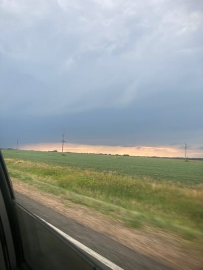 This media file depicts a rural landscape with a distinct weather phenomenon. The scene is divided horizontally, with the upper portion dominated by a gradient of sky, transitioning from a light, overcast blue on the left to a more dramatic, stormy grey. This grey band suggests an approaching or receding storm front. Below the sky, a flat horizon separates it from the land.
The land itself is primarily composed of a vibrant green field, likely agricultural, stretching as far as the eye can see. The field appears to be cultivated in neat rows. A dirt or gravel path runs alongside the field, and the right side of the frame shows a blurred section of what appears to be the edge of a vehicle, indicating the image was taken from inside a moving car.
Several man-made structures, likely power line poles, are visible dotting the horizon across the field. The lighting suggests it is either early morning or late afternoon, as the light is diffused and there are hints of warmer tones near the horizon where the sky meets the land. The overall mood is one of vastness and the raw power of nature, with the impending storm adding a sense of drama to the otherwise serene rural setting. There is no visible text in the media file.