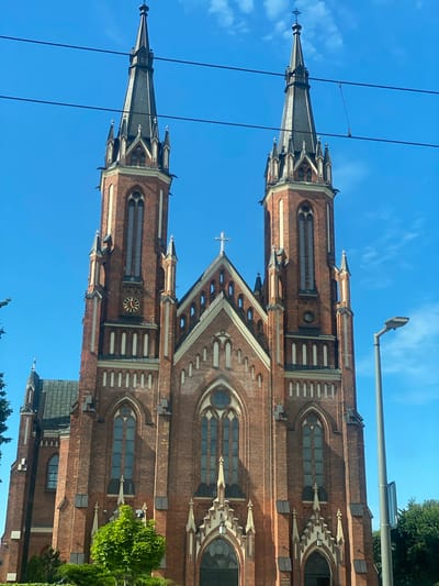 The image shows a view of a red brick church with two tall spires against a clear blue sky. The church architecture is Gothic Revival, characterized by pointed arches, intricate stonework, and steep spires. One of the spires is topped with a cross. There is a clock on one of the church towers, with Roman numerals visible. Wires from an overhead power line are visible in the foreground, obscuring parts of the church and sky. A street light is also visible above the church. Some green trees and foliage are visible on the right side of the image, suggesting an outdoor setting. The image appears to have been taken during the daytime, under sunny weather conditions. The orientation of the image is portrait mode, and the scene is rotated 90 degrees counter-clockwise. Based on the provided location context, the church is likely located in Pabianice, Poland. No people are visible in the image.