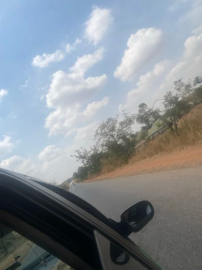 The image depicts a daytime scene on a rural or semi-urban road in Tataura, Nigeria, viewed from inside a dark-colored vehicle. The lower left and center of the frame show the car's window frame, a small portion of the dashboard, and the passenger-side mirror. The paved asphalt road, appearing two-lane, extends into the hazy distance and curves slightly to the left. A reddish-brown dirt shoulder borders the right side of the road, transitioning into dry, light-brown vegetation, including grasses and sparse trees with minimal foliage. A structure with a light-colored, possibly green, roof is partially visible in the mid-ground, nestled amongst the dry trees and brush on the right side of the road. Faint outlines of other vehicles are discernible further down the road. The upper two-thirds of the image display a bright blue sky with scattered white cumulus clouds. No individuals are explicitly visible.