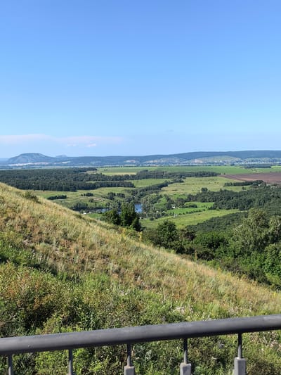 The image shows a panoramic view of a rural landscape under a clear blue sky. In the foreground, a grassy slope descends, with a metal railing along the bottom edge. The midground features a vast expanse of green fields, interspersed with patches of trees and a winding river or lake. In the distance, a range of rolling hills forms the horizon. The overall scene depicts a serene and natural environment, possibly in the vicinity of Akhmerovo, Russia, given the provided context. There are no people or explicit actions depicted in the image.