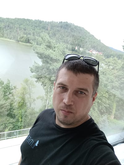 A man with short brown hair and a beard, wearing a black t-shirt with a blue logo and sunglasses on his head, poses for a selfie. He is indoors, with a large window behind him that looks out onto a lush green landscape. In the background, a body of water, likely a lake, is visible, bordered by dense, tree-covered hills. Small houses with red roofs are scattered among the trees on the far side of the lake. The overall impression is a scenic view from a balcony or room overlooking the natural beauty of the area. The location appears to be Doksy, Czechia, known for its picturesque lake and surrounding forests.