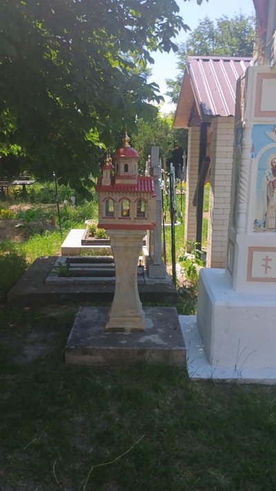 In Mitoc, Moldova, this image captures a serene cemetery scene under a bright sky. The foreground is dominated by a small, intricately designed model of a church, adorned with red roofs and golden domes, standing on a pedestal. This miniature edifice is placed on a concrete base amidst lush green grass and scattered fallen leaves. Surrounding the model, several gravestones and mausoleums are visible. To the right, a larger, more elaborate mausoleum is partially captured, featuring a white structure with painted religious imagery. The background reveals a dense canopy of green trees, casting dappled shadows, and a clear blue sky. The overall atmosphere is one of peaceful solemnity, typical of a cemetery in a rural or semi-rural setting.