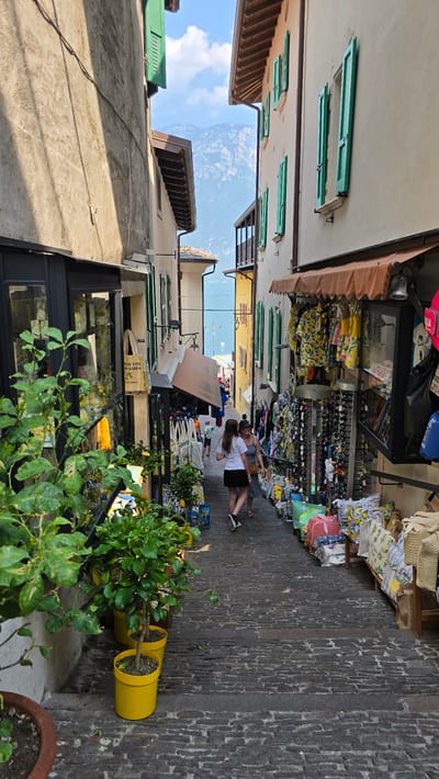 The image depicts a narrow, stone-paved street in Limone sul Garda, Italy, likely during the daytime on a sunny day given the shadows and bright lighting. The scene is characterized by a row of shops on one side, displaying a variety of goods. These include clothing, hats, and souvenirs, many featuring lemon motifs, consistent with the location's name and known produce. On the other side of the street, buildings with green shutters and tiled roofs are visible, with glimpses of a blue sky and distant mountains suggesting a Mediterranean landscape.

In the foreground, lush green plants in bright yellow pots add a touch of vibrant color to the scene. Further down the street, two women are walking, one with dark hair and the other with lighter hair, appearing to be engaged in conversation or browsing the shops. A few other people are also present in the distance, contributing to a relaxed, leisurely atmosphere. The overall impression is one of a charming, picturesque European town, likely a popular tourist destination.

Visible text includes "ALOE VITA DI LIMONE" on a yellow sign, reinforcing the local connection to aloe and lemons. The composition, with the street receding into the distance between the buildings, creates a sense of depth and invitation to explore further.