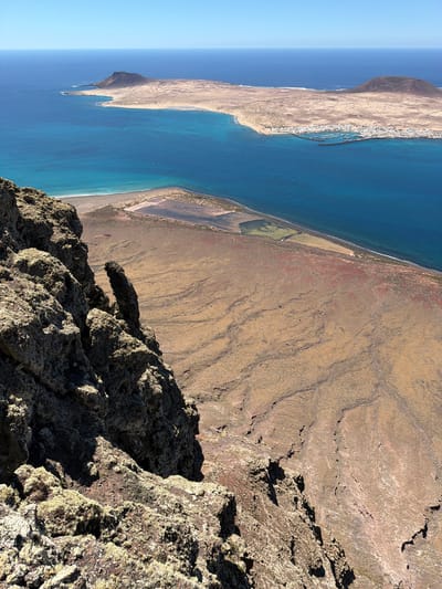 This striking landscape shot, taken from a high vantage point, offers a panoramic view of an arid coastal environment under a clear blue sky. The foreground is dominated by rough, dark volcanic rock formations, some appearing to be covered with light-colored lichen, casting deep shadows that indicate a sunny day, likely mid-morning or late afternoon.

Beyond the immediate rocky precipice, a vast expanse of reddish-brown and tan volcanic terrain stretches towards the sea, marked by subtle patterns suggesting ancient lava flows or erosion. Closer to the coastline, rectangular, man-made structures, possibly salt pans or agricultural plots, are visible near a narrow strip of sandy beach.

The vibrant turquoise and deep blue waters of the ocean meet this barren land. Along the distant coast, a town with numerous white buildings clusters around a harbor or marina, where several boats can be seen docked. Further out, a distinct landmass, likely an island or a prominent headland, rises from the ocean, characterized by its arid, undulating hills.

The image captures the rugged beauty typical of volcanic islands. Given the context, this view is likely from a point in or near Haría, Spain, offering a classic vista of the Lanzarote landscape. There is no visible text in the image.