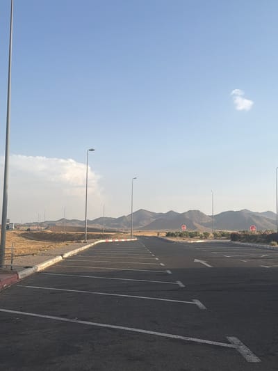The image depicts an empty parking lot on the edge of Jbilate, Morocco. A backdrop of arid mountains is visible in the distance under a hazy sky with a few scattered clouds. The parking lot's asphalt is marked with white lines defining parking spaces and directional arrows. Along the edge of the lot, there's a curb painted with a red and white pattern, bordering a strip of dry vegetation. A series of tall, slender lampposts are spaced along this edge, stretching into the distance. Two red stop signs are visible in the far distance by the mountain and the edge of the parking lot.