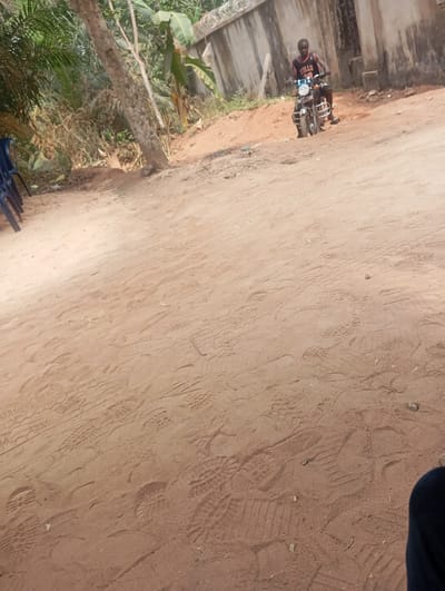 The scene is set outdoors in Okpatu-Ukehe, Nigeria, under bright daylight. The foreground is dominated by reddish-brown earth, extensively marked with various shoe and boot prints. In the mid-ground, a male subject is seated on a dark-colored motorcycle on a slightly elevated dirt path. He is wearing a dark sleeveless top emblazoned with "BULLS" text. His feet are on the ground, indicating the motorcycle is stationary or at rest. The motorcycle's headlight emits a bluish glow. Behind the subject and motorcycle, a concrete wall topped with barbed wire or razor wire spans horizontally, separating the foreground from a dense backdrop of green vegetation, including identifiable banana plants. A structure with a dark aperture, possibly a doorway, is visible beyond the wall to the right of the motorcycle. On the far left of the foreground, two blue plastic chairs are partially visible, stacked and resting against an unseen object amidst sparse vegetation. The lower right corner of the image shows a portion of dark fabric, consistent with a person's leg or knee, suggesting the perspective of the observer.
