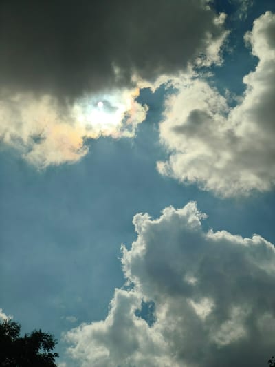The image captures a sky with scattered clouds, and a portion of the sun is visible through a gap in the clouds, creating a bright halo effect with hints of rainbow colors. The sky is a mixture of blue and gray tones, with some clouds appearing dark and heavy, while others are bright and fluffy. In the lower left corner, the dark silhouette of tree branches is visible, suggesting an outdoor scene. The image does not depict any specific location within the city of Nanguan District, China, nor does it contain any people, subjects, objects, or actions. The scene is a natural environment focused on the atmospheric conditions.