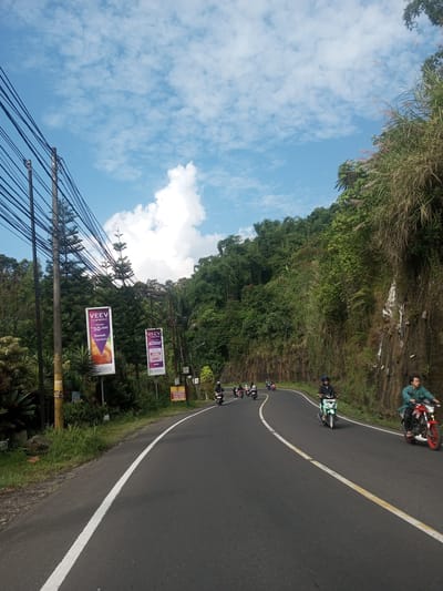 The image captures a scene on a winding road, possibly in a rural area, likely in Southeast Asia. Several motorcyclists are riding on the road, with two prominent riders in the foreground. The road is paved, with white and yellow lines indicating lane separation.
On the left side of the road, there are trees, lush vegetation, and utility poles with numerous wires. Two advertising billboards are visible, promoting the product VEEV, displaying a price of 50,000 and the phrase "Ramah di kantong." One billboard also mentions "SRC."
The right side of the road features a rocky hillside covered in dense green vegetation.
The sky is partly cloudy, with patches of blue visible. The overall setting suggests a tropical or subtropical environment. No specific location in Khe Luang, Laos, can be determined from the image alone.