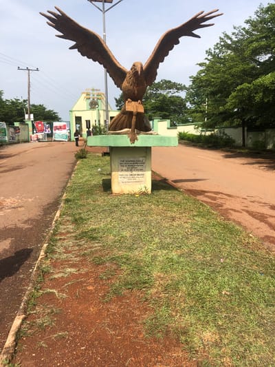 The image depicts a large bronze sculpture of an eagle with its wings spread wide, perched on a book, which rests on a green pedestal. The pedestal has an inscription that reads, "THE SOARING EAGLE, A SYMBOL OF THE KING OF LIMITLESS QUEST OF MAN TO ASCEND. THIS SCULPTURE PRESENTED AT THIS WAS UNVEILED ON THE 3RD DAY OF JULY, 2019 BY PROFESSOR CHINEDUM NWAJIUBA VICE CHANCELLOR, AUGUSTINE UNIVERSITY, ILARA-AKOKO. SCULPTOR: EPHRAIM KELECHI OGUANYI."

Behind the sculpture, a green and yellow building with arched entrances is visible, possibly an entrance gate to an institution. Several posters are affixed to a green gate to the left of the sculpture. Two individuals, one in dark clothing and another in lighter clothing, are walking away from the viewer in the direction of the building. The scene is set on an asphalt road, with a strip of grass and red soil bordering it. Tall trees with lush green foliage are present in the background, and the sky is partly cloudy. The overall impression is that of an outdoor sculpture at the entrance of an educational institution. The location is stated as Eka, Nigeria.
