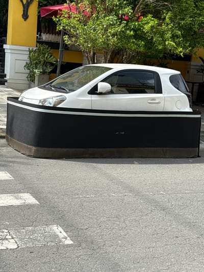 A white two-door hatchback vehicle is stationary on cracked asphalt pavement in An Hải Ward, Vietnam. The car is partially enveloped by a large, dark, curved rectangular structure, appearing as a promotional display or a modified outdoor furnishing. This structure is black with a narrow white horizontal stripe near its top edge and a brown base, obscuring the car's lower half and wheels. In the background, a building with a yellow facade, white trim, and a red awning is visible, accompanied by lush green trees and potted plants, indicating an urban commercial setting. The foreground asphalt features worn, faded white crosswalk markings. The scene is well-lit by daylight; no individuals or dynamic actions are depicted.