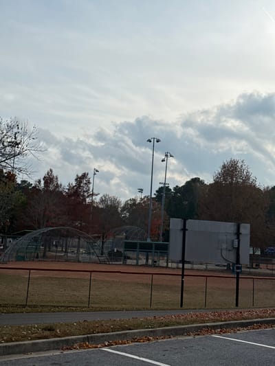 An outdoor sports field on a cloudy day. Fences enclose the field, and tall light poles stand along the perimeter. Trees with autumn foliage line the edge of the field. A gray building or structure is present. An asphalt surface with white lines borders the field. The sky is filled with gray and white clouds.