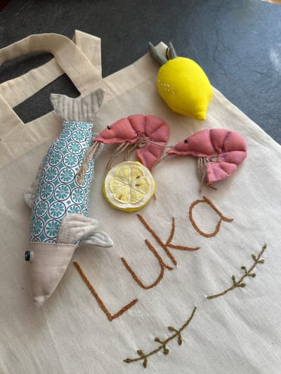 A close-up shot shows a canvas tote bag on a dark, textured surface. Embroidered onto the bag in brown thread are the letters "LUKA" and a decorative leafy vine. To the left of the embroidery, there are three handmade fabric decorative items: a fish with a blue and white circular pattern and gray fins, two pink shrimp, and a slice of lemon. A yellow fabric lemon with green leaves is positioned above the shrimp. The background suggests an indoor setting.