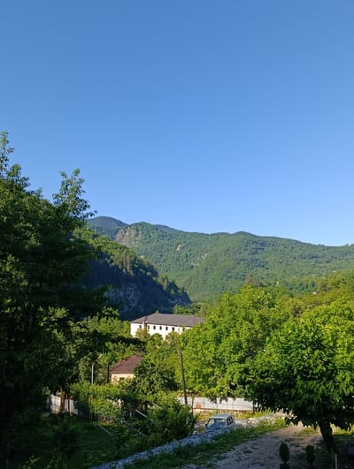 The image depicts a serene landscape with lush green mountains under a clear blue sky. In the foreground, several trees frame the scene, with a footpath leading into the distance. A small residential building with a brown roof is visible, surrounded by greenery and a fence. Further back, a larger, light-colored building with a dark roof stands amidst the trees. A light-colored pickup truck is parked on the path. The scene appears to be a rural residential area in the city of Achara, Georgia, characterized by its natural beauty and tranquil atmosphere.