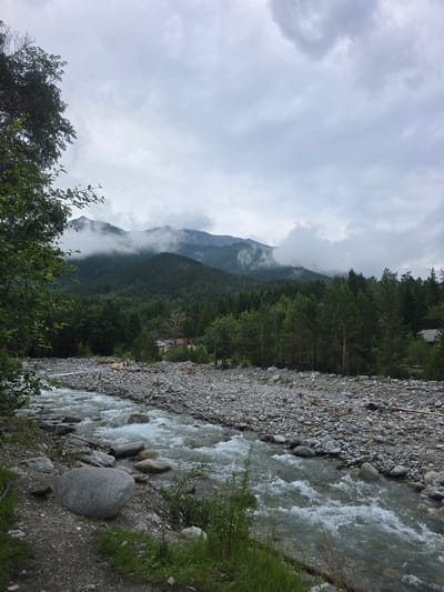 This is an eye-level, outdoor shot of a rocky riverbed in Arshan, Russia. The river flows from the bottom right of the frame towards the top left, with whitewater rapids visible. The riverbanks are lined with numerous grey and brown stones of varying sizes, interspersed with patches of green grass and small bushes. In the background, a dense forest of green pine trees covers rolling hills. The forest gives way to a mountain range in the distance, with its peaks shrouded in white clouds. The sky above is overcast and a pale grey. A few small, rustic buildings with red roofs are nestled among the trees in the mid-ground. Several people are visible on the riverbank in the distance, some appearing to stand on a log bridge.
