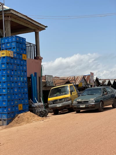 The image shows a street scene in Tangu, Nigeria, under a bright, partly cloudy sky. To the left, a large stack of blue plastic crates, possibly for beverages, is piled high against a building with a corrugated metal roof supported by pillars. Some yellow crates are visible at the top of the stack, one labeled "STAR." Next to the crates, a pile of sand and two metal trolleys are present.

In the center of the image, two vehicles are parked on a dirt road. A yellow truck is parked in front of a dark green Volkswagen Golf. The truck has a license plate that reads "JJR 301XA." The Golf, which appears older and shows some rust, has a license plate that reads "MKA-8036R." Behind the vehicles, several market stalls with corrugated metal roofs are visible. A framed poster featuring two men is displayed on the wall of one of the stalls. Blue pipes are leaning against the building next to the stacked crates. The overall environment suggests an outdoor market or commercial area.
