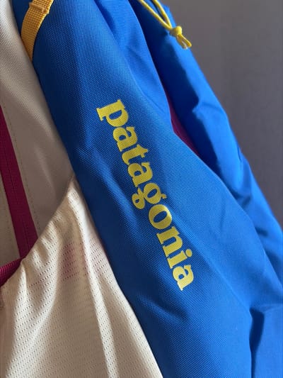 A close-up, angled shot displays sections of multi-colored technical apparel. The primary subject is a vibrant blue fabric, characteristic of an outdoor jacket or similar garment, with the brand name "patagonia" printed vertically in bright yellow, lowercase letters on its surface. To the immediate left, a section of cream-colored mesh fabric is visible, separated from the blue material by a thin, vertical magenta strip. A yellow fabric strap or trim is present near the top left edge, attaching to the blue garment, while a yellow knotted drawstring is observed on the blue fabric in the upper right. The blue material exhibits a structured, finely textured weave, and the cream fabric features a distinct mesh pattern. The background is an indistinct, plain light grey surface, suggesting an interior setting. No individuals or specific actions are depicted. The specific location in the city of Krasnaya Zarya, Russia, cannot be determined from the visual evidence provided in the image.
