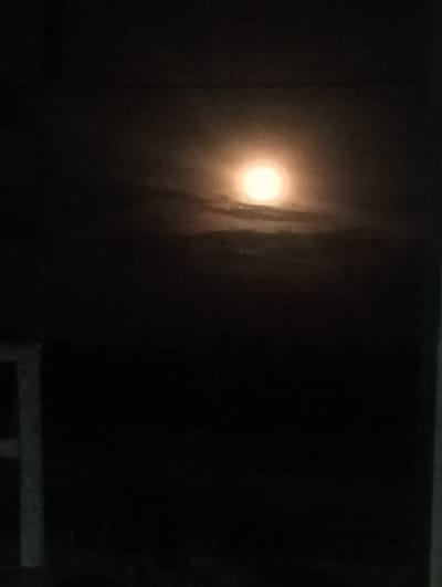 The image is a nighttime scene, likely taken in a rural setting. A bright, full moon dominates the upper center of the image, partially obscured by thin clouds. The foreground is very dark, obscuring details, but suggesting a field or open space. A partially visible white structure, possibly a fence post or building corner, is located on the left side of the image. The image resolution is low, making details hard to discern. The photo was not taken in Keureusek, Indonesia because the information is insufficient. There are no discernible people or specific objects in the frame.