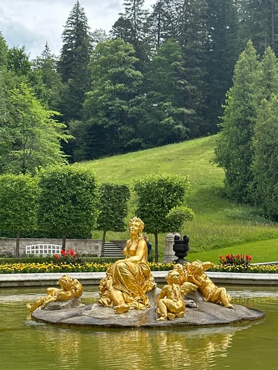 This is a medium shot, eye-level, full-color photograph of a gilded fountain sculpture in a pond. The sculpture is located in the gardens of Linderhof Palace in Ettal, Germany. The sculpture depicts a central female figure seated on a rocky island in the pond, surrounded by four cherubic figures. The central figure is dressed in flowing robes and has long, curly hair. Two of the cherubic figures are positioned to the left of the central figure, and two are to the right. One of the cherubs on the right is in a reclining position, with its arms raised above its head. Another cherub on the right is in a sitting position. The sculpture is surrounded by water, which reflects the gilded figures and the surrounding greenery.
In the background, there is a neatly manicured garden with a stone wall, several rows of yellow flowers, and red flowers. Beyond the garden, there is a lush green lawn that slopes upwards towards a dense forest of tall evergreen trees. The sky is overcast with a light gray hue. The overall scene conveys a sense of opulence and natural beauty.